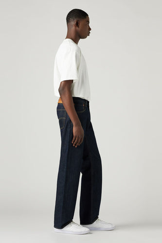 Man staat zijdelings, witte T-shirt, donkere jeans, witte sneakers, lichte achtergrond.
