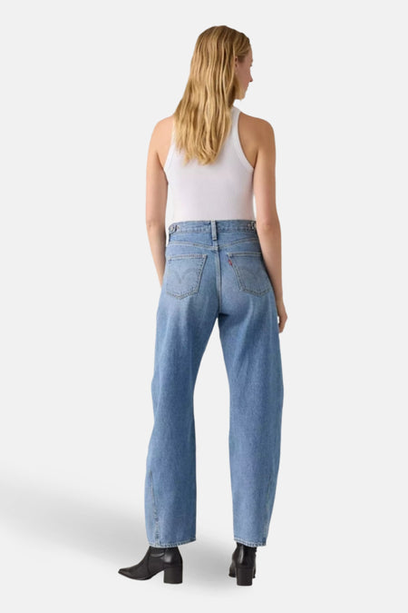 Achteraanbeeld van een vrouw in een witte tanktop, wijde blauwe jeans en zwarte laarsjes.