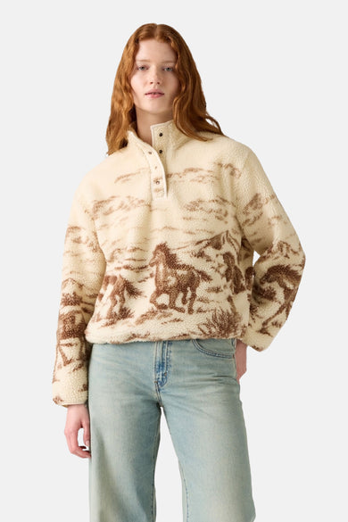 Een vrouw draagt een beige fleece trui met een paardenprint en blauwe jeans.