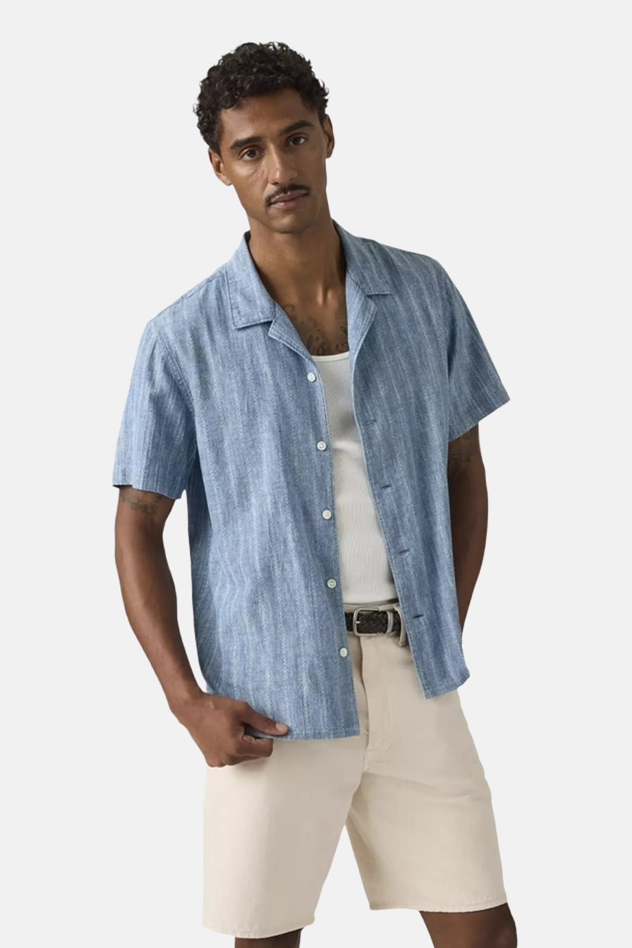 Chemise bleue à manches courtes de Levi's®, avec une fine texture verticale, ouverte sur un débardeur blanc et un pantalon de couleur crème.
