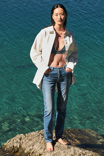 Jean droit en denim bleu clair de MAC, porté avec un chemisier en lin ouvert et une brassière en denim sur un rocher au bord de l'eau.
