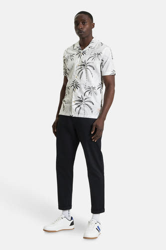 Chemise blanche à manches courtes de BEN SHERMAN, avec un imprimé palmier et associée à un pantalon noir et des baskets blanches.
