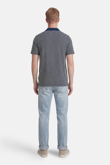 Blauw-wit gestreepte polo van BEN SHERMAN, met een donkerblauwe kraag, gedragen met lichte jeans en suède schoenen, gezien van achteren.
