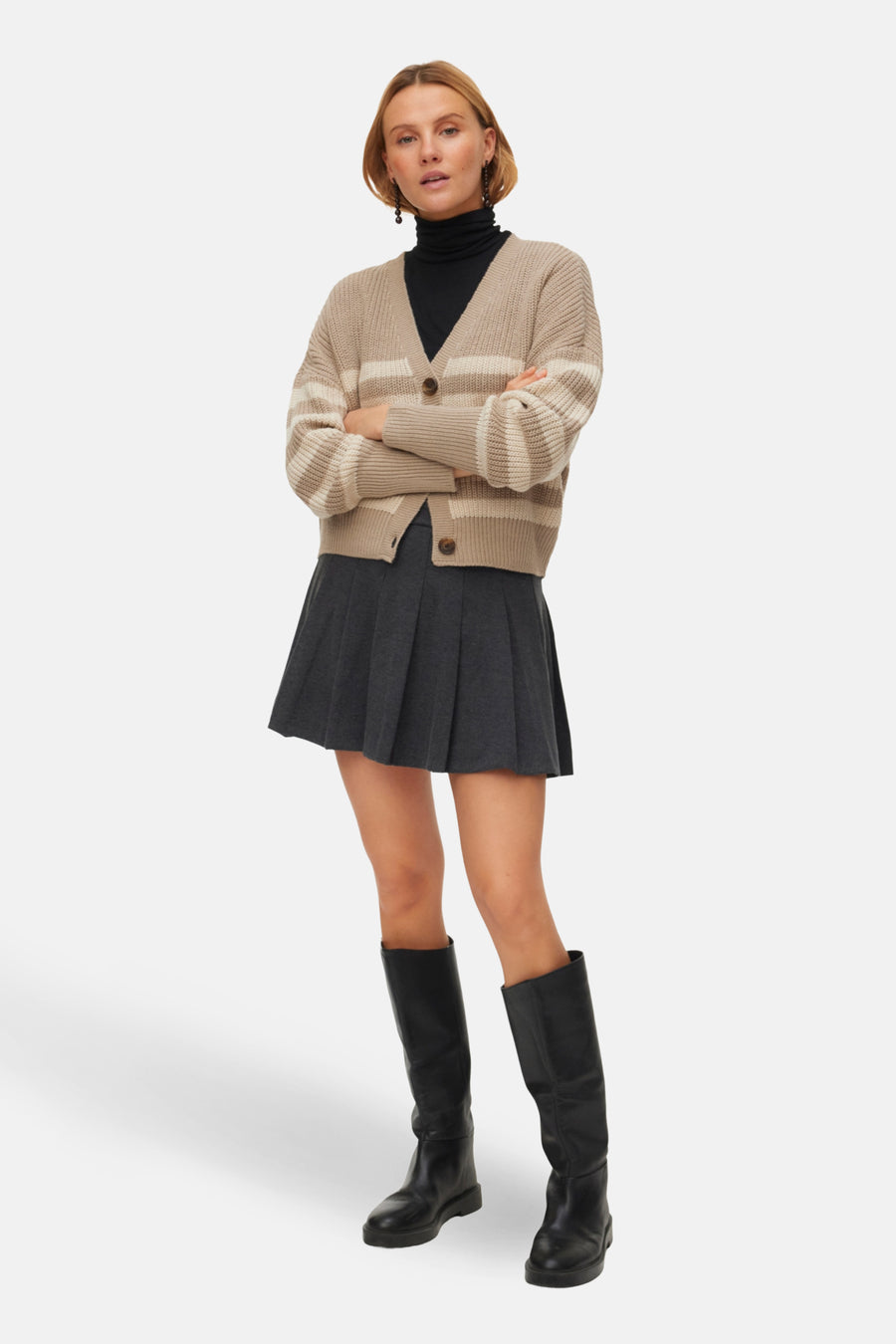 Vrouw in geribbelde beige cardigan, zwarte coltrui, grijze plooirok en zwarte laarzen.
