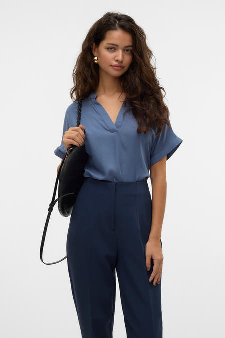 Blouse bleue à manches courtes de VERO MODA, avec un col en V et portée avec un pantalon bleu marine et une sac noir.