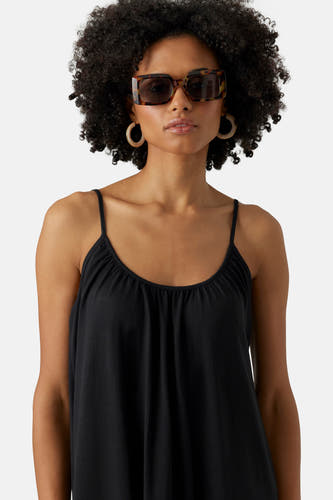 Robe noire sans manches de VERO MODA, avec des bretelles spaghetti, portée avec des boucles d'oreilles dorées et un chapeau de soleil.