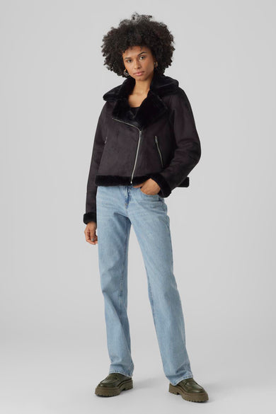 Manteau court noir de VERO MODA, avec une fermeture éclair asymétrique et un col teddy, associé à des jeans bleus et des chaussures vertes.