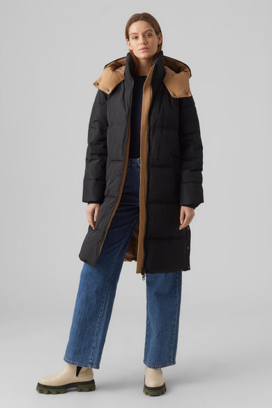 Manteau long noir de Vero Moda, avec capuche et jeans bleus et baskets beige.