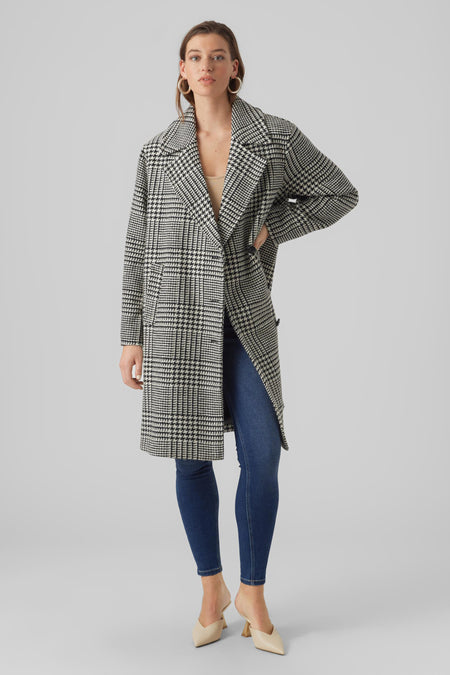 Manteau long noir de Vero Moda, avec un motif à carreaux, porté avec des jeans et des baskets.