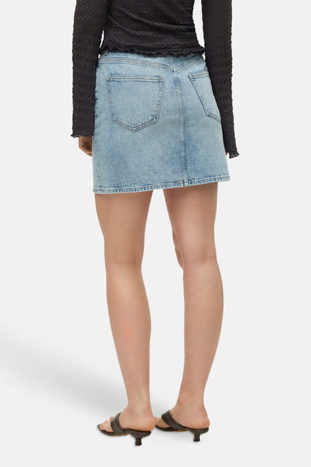 Jupe courte en denim bleu clair de VERO MODA® avec deux poches arrière, vue de dos, portée avec un haut noir et des talons.