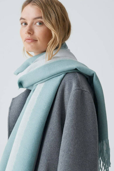 Blauwe wintersjaal van OPUS, met een lichtblauwe en witte streep over een grijze jas.
