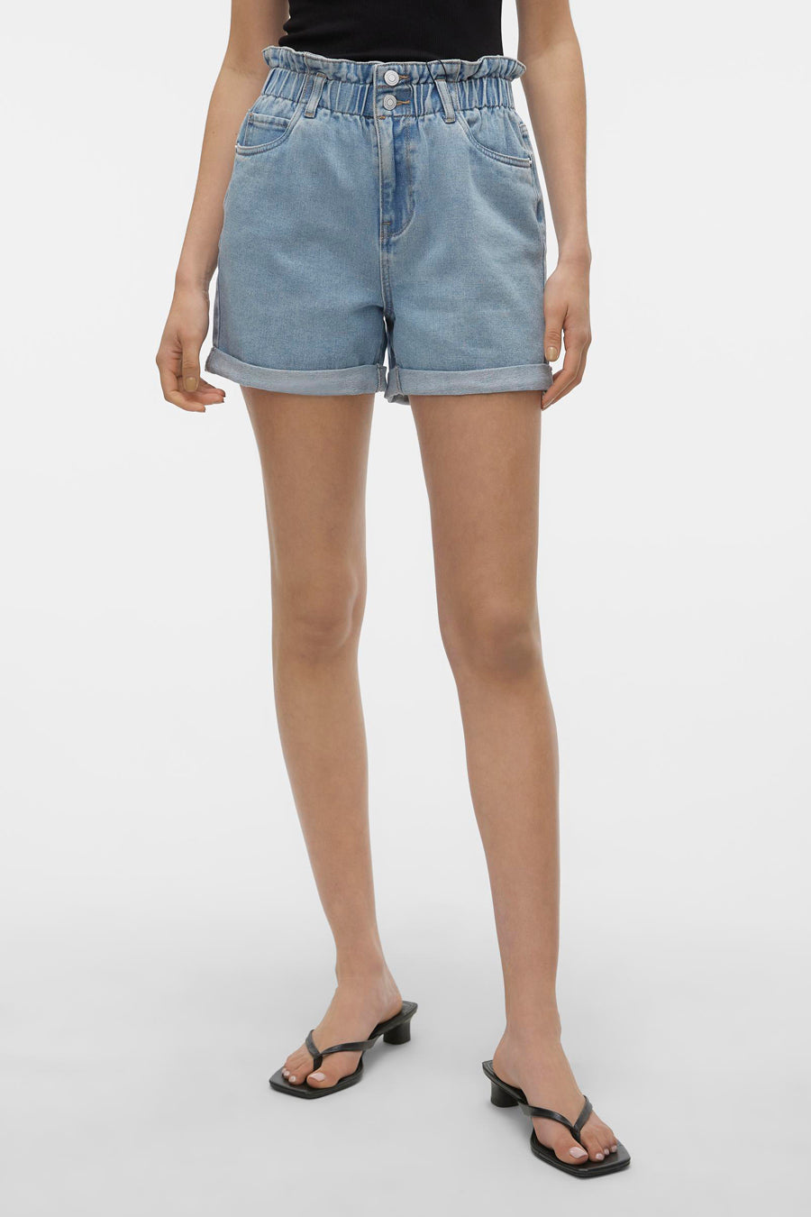 Short en jean bleu de VERO MODA® avec taille paperbag élastique haute, ourlets retroussés et poches arrière.Vue de dos, portée avec un haut noir et des mules.