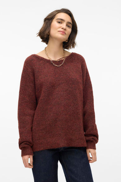 Vrouw met roodbruine trui en gouden ketting, casual pose voor witte achtergrond.
