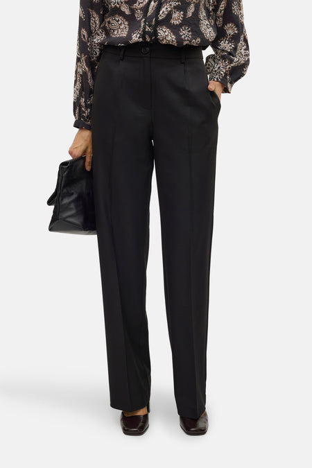 Pantalon noir de VERO MODA, avec des jambes droites, porté avec une blouse imprimée et un sac à main en cuir.