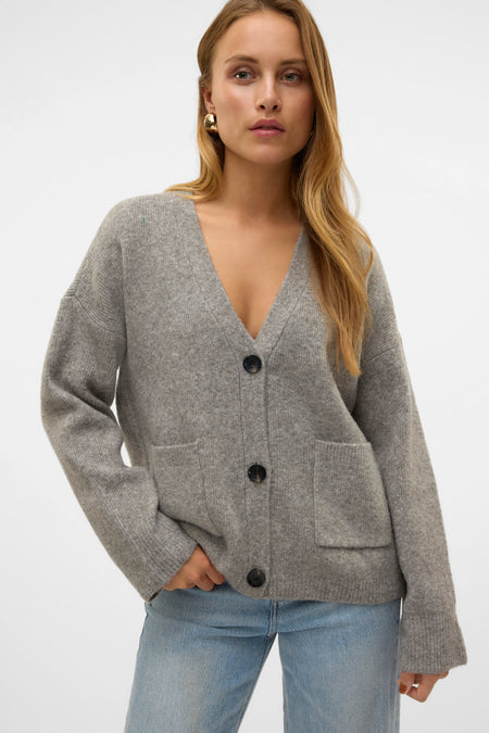 Veste grise de VERO MODA, avec des manches longues, une fermeture à boutons et des poches brodées, portée sur un pantalon de cuir bleu.