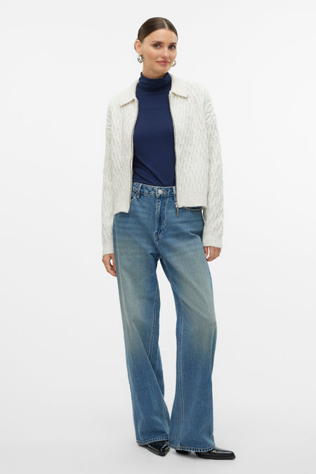Ecru gilet van VERO MODA met kabelpatroon, gedragen over een donkerblauw coltrui en wijde jeans.
