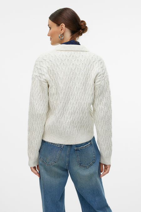 Ecru gilet van Vero Moda, met een kabelpatroon, gedragen over een blauw shirt en een spijkerbroek.

