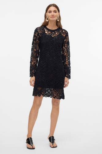 Zwarte kanten jurk met lange mouwen en gouden oorbellen, staand tegen witte achtergrond.