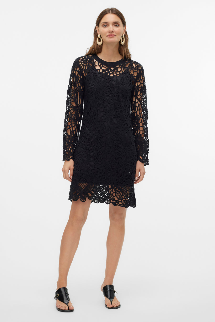 Zwarte kanten jurk met lange mouwen en gouden oorbellen, staand tegen witte achtergrond.