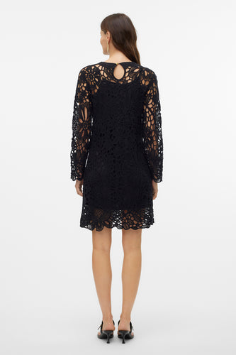 Achterkant van een vrouw in een zwarte, lange mouwen kanten jurk en zwarte hakken, staand voor een witte achtergrond.