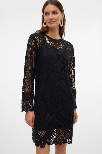 Een vrouw in een zwarte, gehaakte jurk met lange mouwen en gouden oorbellen, tegen een witte achtergrond.