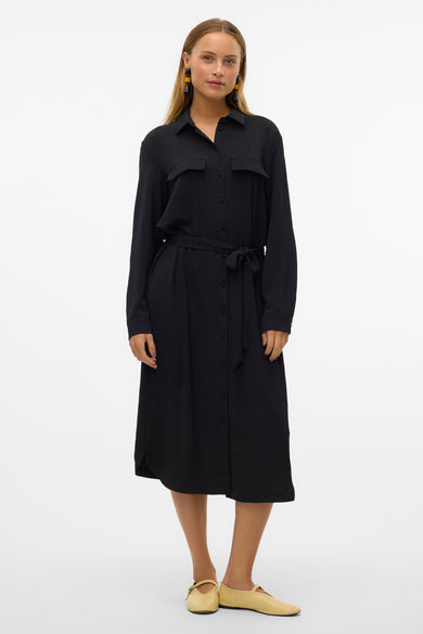 Robe noire à longues manches de VERO MODA®, avec fermeture à boutons et ceinture à la taille, portée avec des chaussures plates.