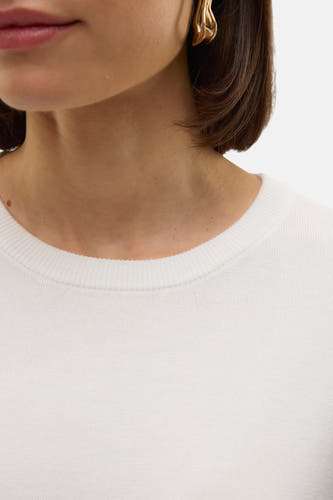 Pull blanc à col rond de VERO MODA, gros plan avec boucles d'oreilles dorées et cheveux châtain foncé.