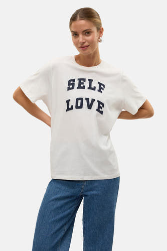 T-shirt blanc à manches courtes de VERO MODA® avec le texte "SELF LOVE" en noir, associé à des jeans bleus.