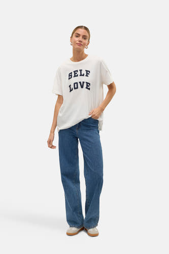 Wit T-shirt met korte mouwen van VERO MODA®, met de tekst "SELF LOVE" in het zwart, gecombineerd met blauwe jeans.
