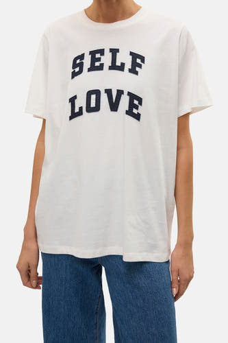 T-shirt blanc à manches courtes de VERO MODA avec du texte bleu marine "SELF LOVE", associé à un jean bleu.