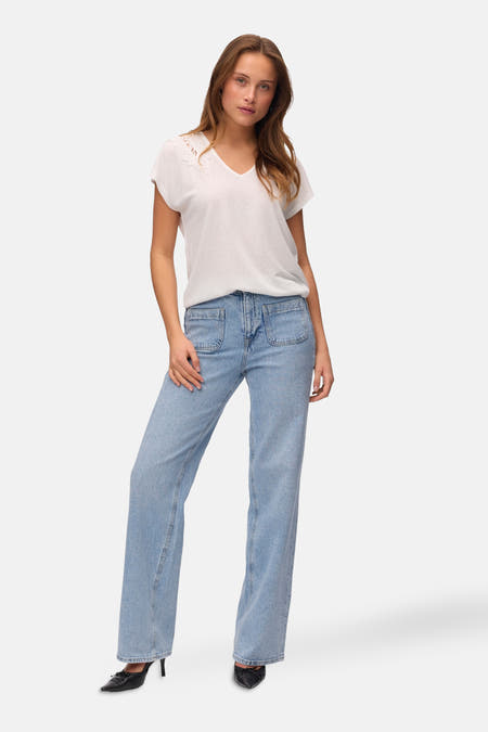 T-shirt blanc à manches courtes de VERO MODA®, avec des détails sur les bords au niveau du col, associé à un jeans bleu et des chaussures noires.