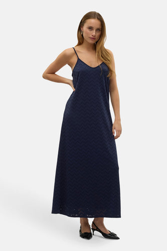 Vrouw in een lange marineblauwe jurk met een V-hals en spaghettibandjes, staand met haar hand op haar heup.