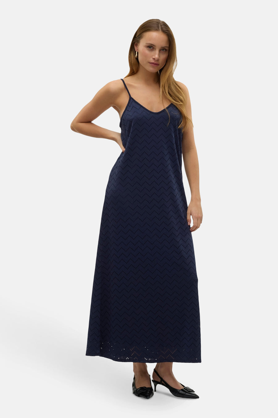 Vrouw in een lange marineblauwe jurk met een V-hals en spaghettibandjes, staand met haar hand op haar heup.