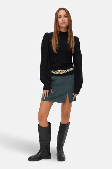 Pull-over noir avec col rond de VERO MODA, manches longues, associé à une jupe verte, une ceinture imprimée zèbre et des bottes noires.