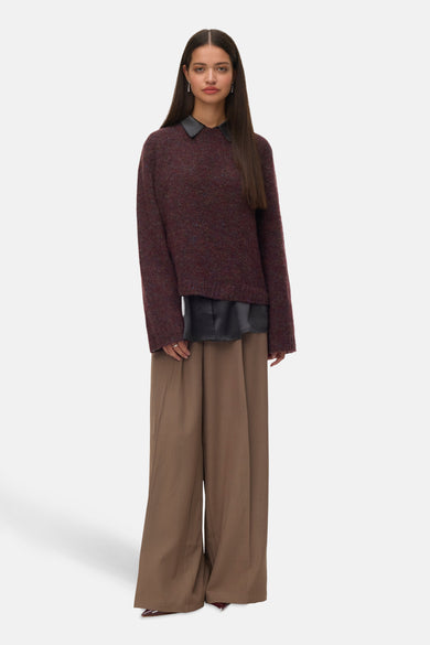 Bordeaux trui met ronde hals van VERO MODA®, gedragen over een zwarte blouse en wijde, bruine pantalon.
