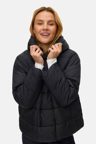 Manteau court noir de VERO MODA, porté avec un chemisier blanc, des jeans bleu marine et des bottes brunes.