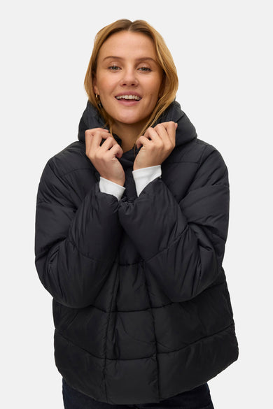 Manteau court noir de VERO MODA, porté avec un chemisier blanc, des jeans bleu marine et des bottes brunes.
