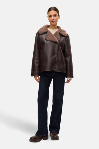 Manteau court brun de VERO MODA, avec un col en fourrure, porté sur une chemise crème, associé à des jeans bleu marine et des bottes brunes.