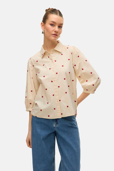 Chemise beige à manches courtes de Vero Moda, avec un motif de cœurs rouges sur toute la surface, associée à des jeans bleus.