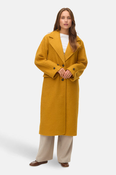 Manteau jaune de VERO MODA avec une longue coupe, porté sur un chemisier blanc et un pantalon beige.