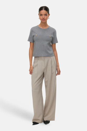 Sweat-shirt gris de VERO MODA, avec des manches courtes et des boutons dorés sur la poitrine, associé à un pantalon beige large et des chaussures noires.