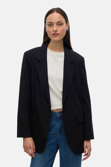 Blazer noir de VERO MODA, avec des manches longues, porté avec un chemisier blanc et des jeans bleus.
