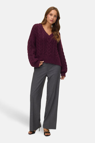 Vrouw in bordeauxrode kabeltrui en grijze pantalon, staand voor een witte achtergrond.