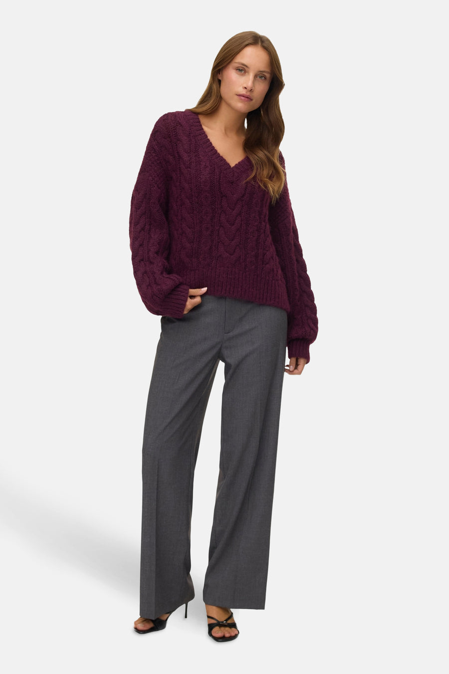 Vrouw in bordeauxrode kabeltrui en grijze pantalon, staand voor een witte achtergrond.