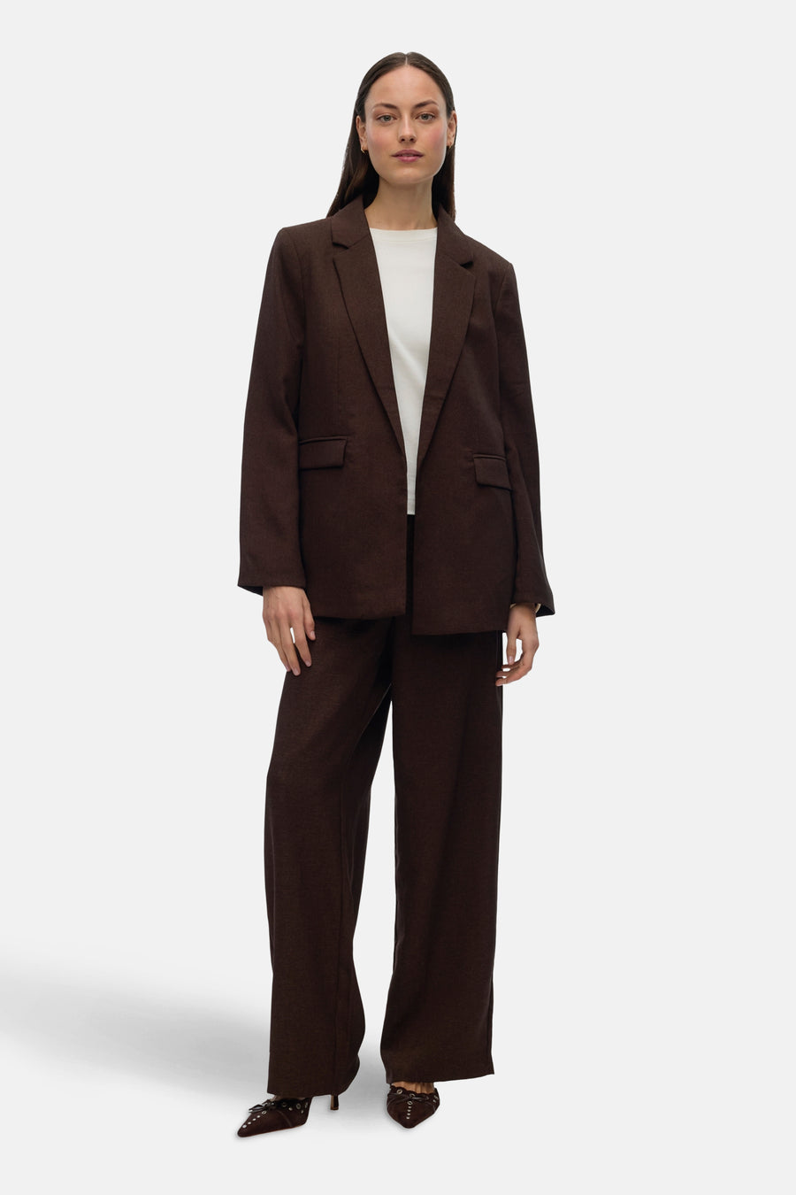 Vrouw in bruin pak met witte top, staand.
