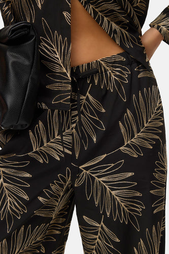 Close-up zwarte broek met gouden bladmotief, zwarte leren tas.