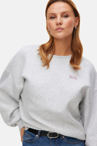 Pull-over gris de VERO MODA®, avec le mot "Faith" brodé, porté avec un jean bleu et une ceinture noire.