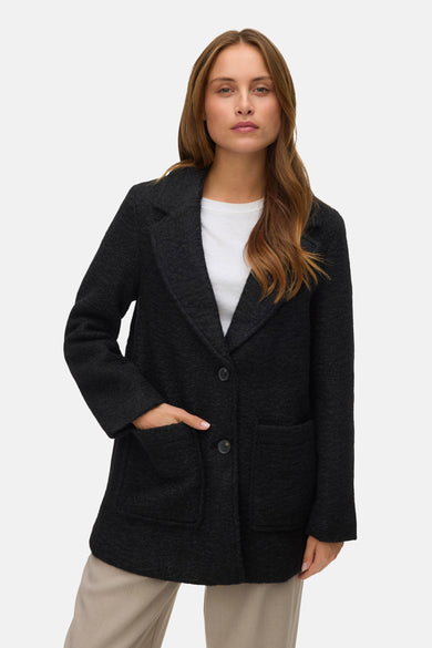 Manteau court noir de VERO MODA avec une texture irrégulière, porté sur un t-shirt blanc et un pantalon beige.