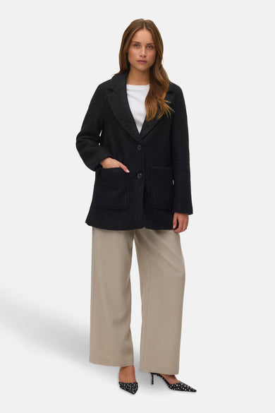 Manteau court noir de VERO MODA, avec des manches longues, une fermeture à boutons et des poches, porté avec un pantalon beige et des escarpins pointus.