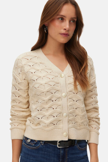 Veste écru de VERO MODA, avec fermeture à boutons et un motif tricoté ajouré, associée à des jeans foncés et des chaussures noires.